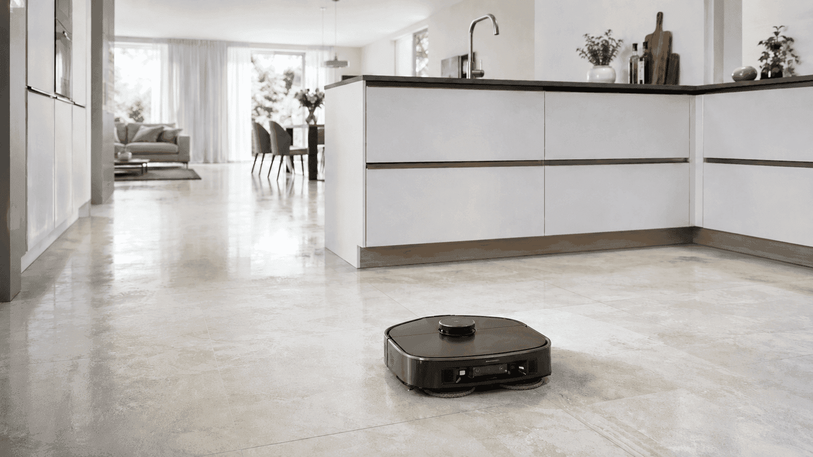 Saug- und Wischroboter fährt auf hellem Küchenboden in moderner offener Küche mit Kücheninsel und Essbereich im Hintergrund.
